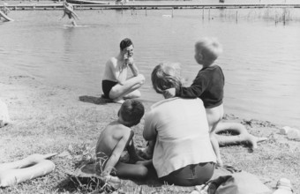Family in the 1950s. A mother and her children sits on the beach while her husband is filming her
