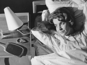 Sleep in the 1960s. A young woman lies comfortably ensconced in a bed. On the bedside table is a