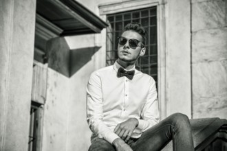 Attractive young man sitting next to walls in city, looking away, wearing sunglasses. Half body