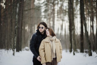 A couple in warm clothing stands in a snowy forest, exuding a serene atmosphere