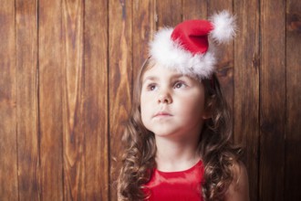 Girl in Santa hat headband sitting over wooden wall, copy space