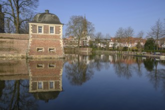 A pavilion at the moat with a clear reflection, surrounded by trees in the park in front of a blue