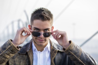Handsome trendy young man standing on a sidewalk wearing sunglasses and a fashionable leather