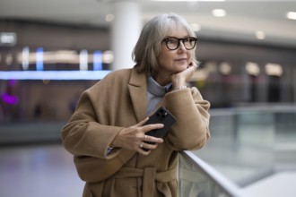 A woman with stylish glasses leans against a railing, thoughtfully holding her smartphone. She