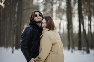 The couple stands happily in a snowy forest, wearing warm clothing and sunglasses