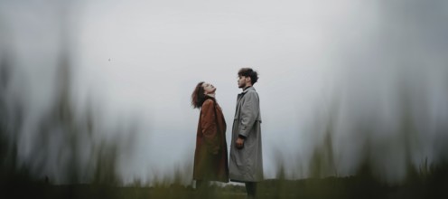 A couple standing in a field, each looking upwards, dressed in long coats with a sense of