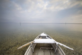 A snow-covered boat on a calm lake with pebbles on the shore under a cloudy sky, Strandbad Hörnle,