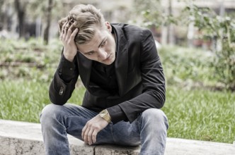 Close up Disappointed Young White Man Sitting at Street Side with Grassy Landscape at the