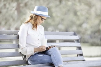 Happy young student with a tablet and a disposable coffee cup sitting on a bench and reading in a