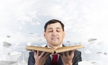 Happy businessman holding open old book. Smiling man in suit standing on downtown background with