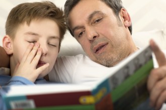 Close-up shot of a father reading a book to the son who is already sleepy and rubbing his eyes
