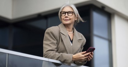 The stylish elderly woman stands elegantly on a balcony, holding her phone and gazing thoughtfully