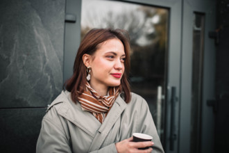 Woman in light coat and striped scarf holding a cup, appearing thoughtful