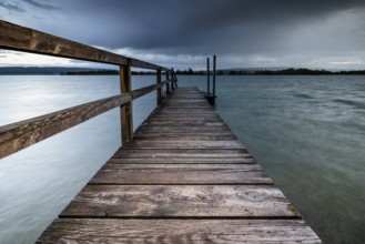 A long wooden footbridge leads into the water of a lake under a dark, cloudy sky, Naturfreundehaus,