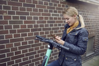 Trendy young blond woman in leather jacket standing at her e-scooter booking a ride, ecological