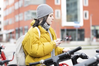 Woman in yellow jacket scrolling on her smartphone next to parked rental bike in the city,