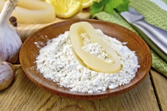 Raw squid rings in flour on a plate, garlic, knife, lemon, napkin on the background of wooden