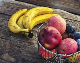 Ripe apples, bananas and plums in metal basket on wooden background