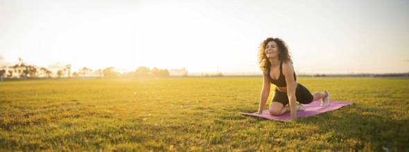 Person smiles while in a yoga pose on a mat, lit by the morning sun, in a field