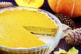 Pumpkin pie in a glass pan on sackcloth, pumpkins, pine cones and maple leaf on the background of