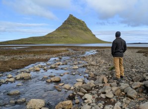 Person standing by a rocky river, gazing at a distinctive mountain under a cloudy sky. Iceland