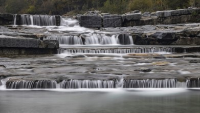 An impressive, multi-levelled waterfall flows over stones in a tranquil natural setting, Val