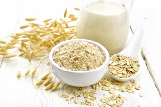 Flour oat in bowl, milk in a jug, oatmeal in spoon, oaten stalks on the background of light wooden