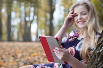 A beautiful girl in an autumn park with a tablet sits near a tree and looks at the camera