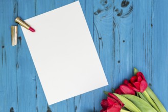 Red tulips, lipstick and a white sheet of paper for your greetings on the background of blue wooden