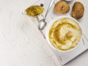 Mashed potatoes in white bowl on white cutting board, pepper and olive oil on white concrete