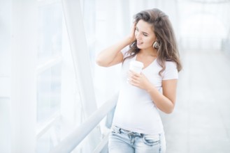 Happy young woman with music headphones standing on the bridge with a takeaway coffee mug, looking