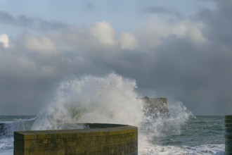 Waves crash against a stone pier wall, with a cloudy sky above and the ocean extending into the