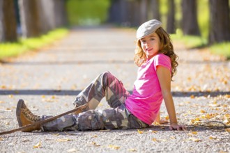 Autumn kid girl with camouflage pants sitting resting in fall trees track with hiker branch stick