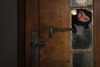 Burglar looks through broken glass door, holds crowbar, gloomy atmosphere, staged photo shoot,