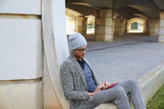 Modern young man with tablet pc touch working under a bridge with wool winter cap