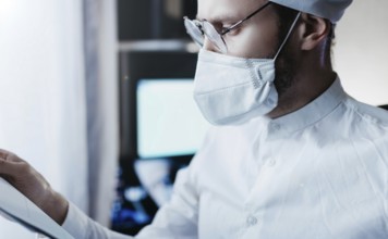 Healthcare worker wearing mask and glasses, carefully examining a document in a clinical setting