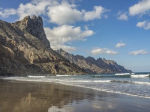 A sandy beach with waves, steep rocks and a clear blue sky with scattered clouds, tenerife, canary