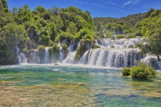 Waterfalls flow into an emerald green river, surrounded by dense forest and a bright blue sky, Krka