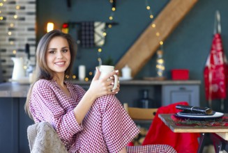 Happy young woman in plaid pajama holding jar smiling at camera in kitchen decorated for Christmas