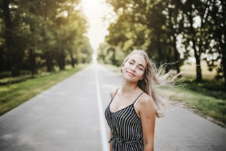 Woman standing on a road surrounded by trees, sunlight filtering through, creating a relaxed summer