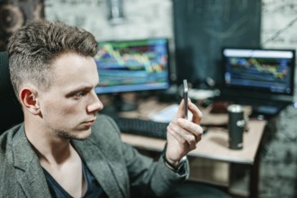 Man examining a smartphone in an office setting with trading monitors behind