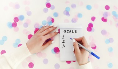 Female hand with pen writing New Year goals in empty notebook on white background with confetti