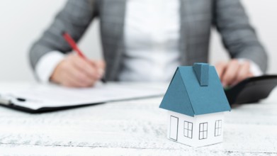 Businesswoman sitting and writing in notebook. Paper house' on desk