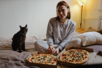 Woman sitting on the bed with pizza boxes and a black cat in a nighttime cozy setting