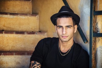 Portrait of handsome man with tattoo on hands sitting on cement stairs while leaning on wall