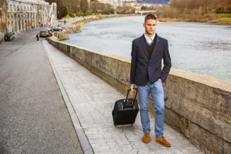 Young man in trendy clothes with baggage standing on pavement near river while looking away