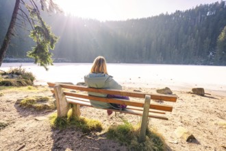 Woman sitting on a bench by a frozen lake, surrounded by a brightly lit, wintry forest,
