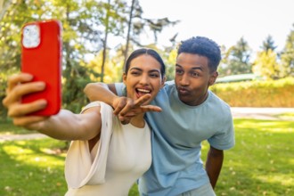 Multi-ethnic couple taking selfie gesturing victory and blowing a kiss in a park