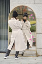 A woman stands outdoors, looking at her reflection in a modern mirror while wearing a coat and a