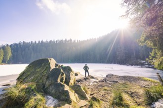Man standing on rocks in front of a frozen lake, surrounded by forest, sunlight illuminates the